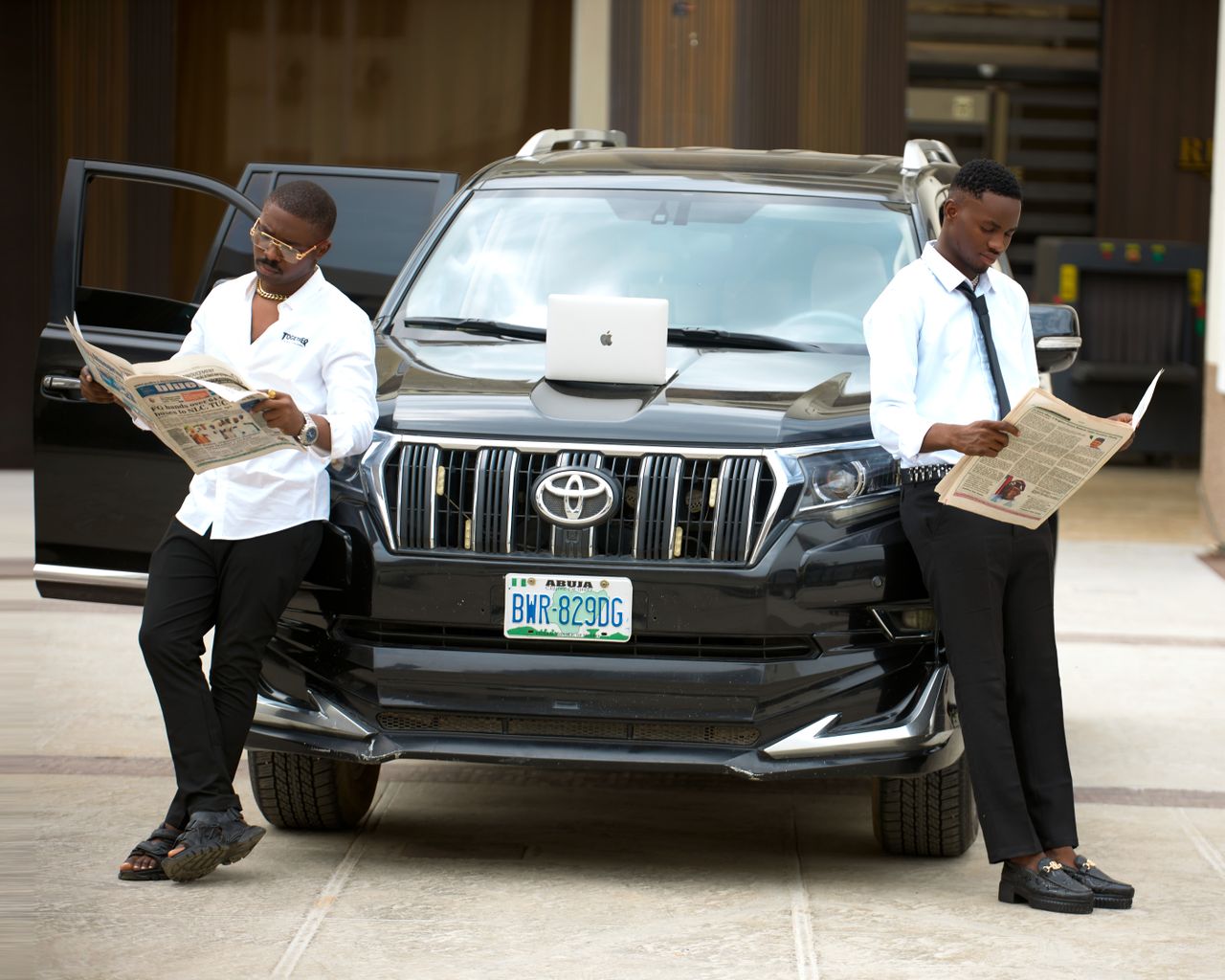 Two men in formal attire reading newspapers by a luxury SUV in an urban setting.